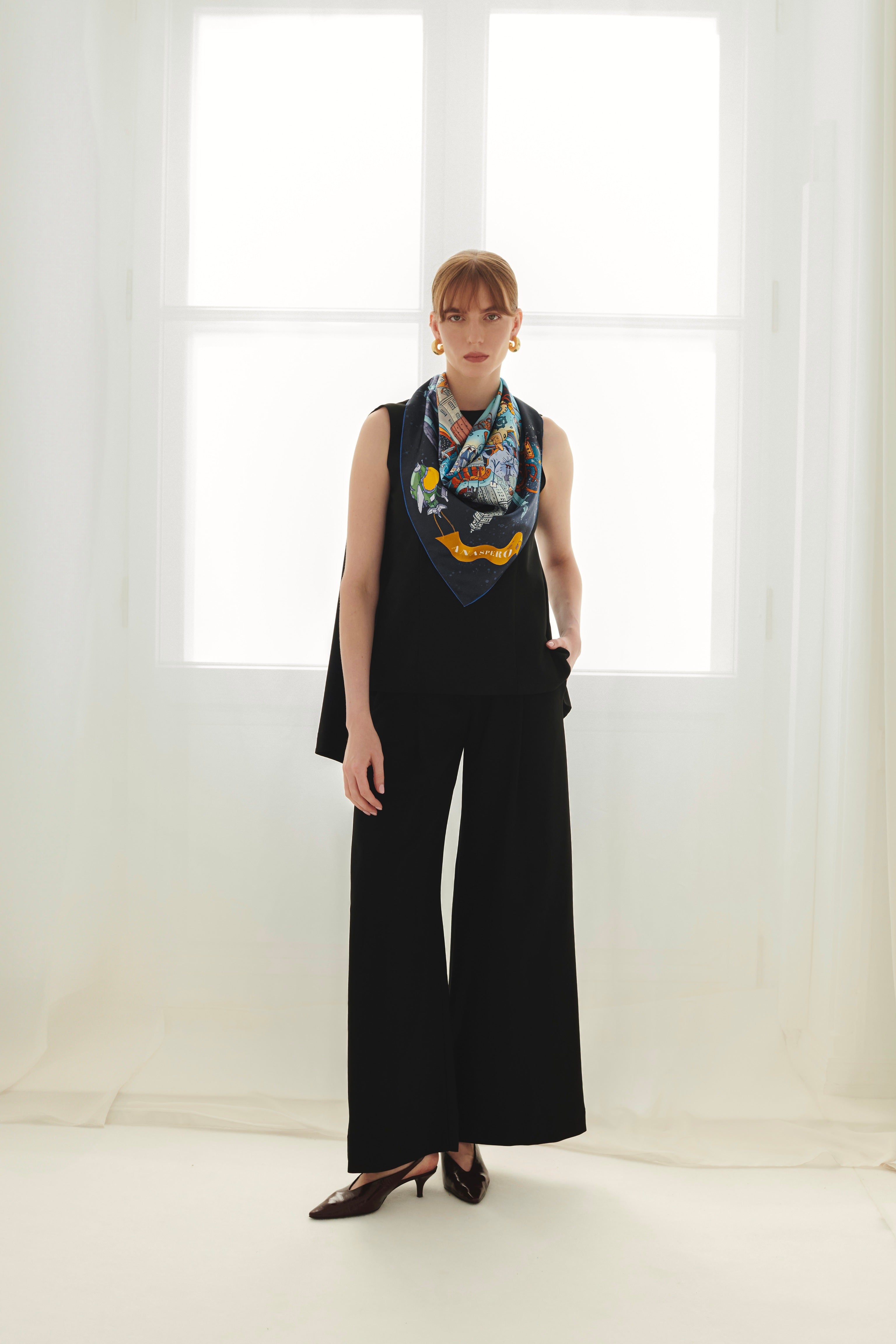 Woman wearing a black sleeveless top and wide-leg pants with a colorful silk scarf in a bright room.