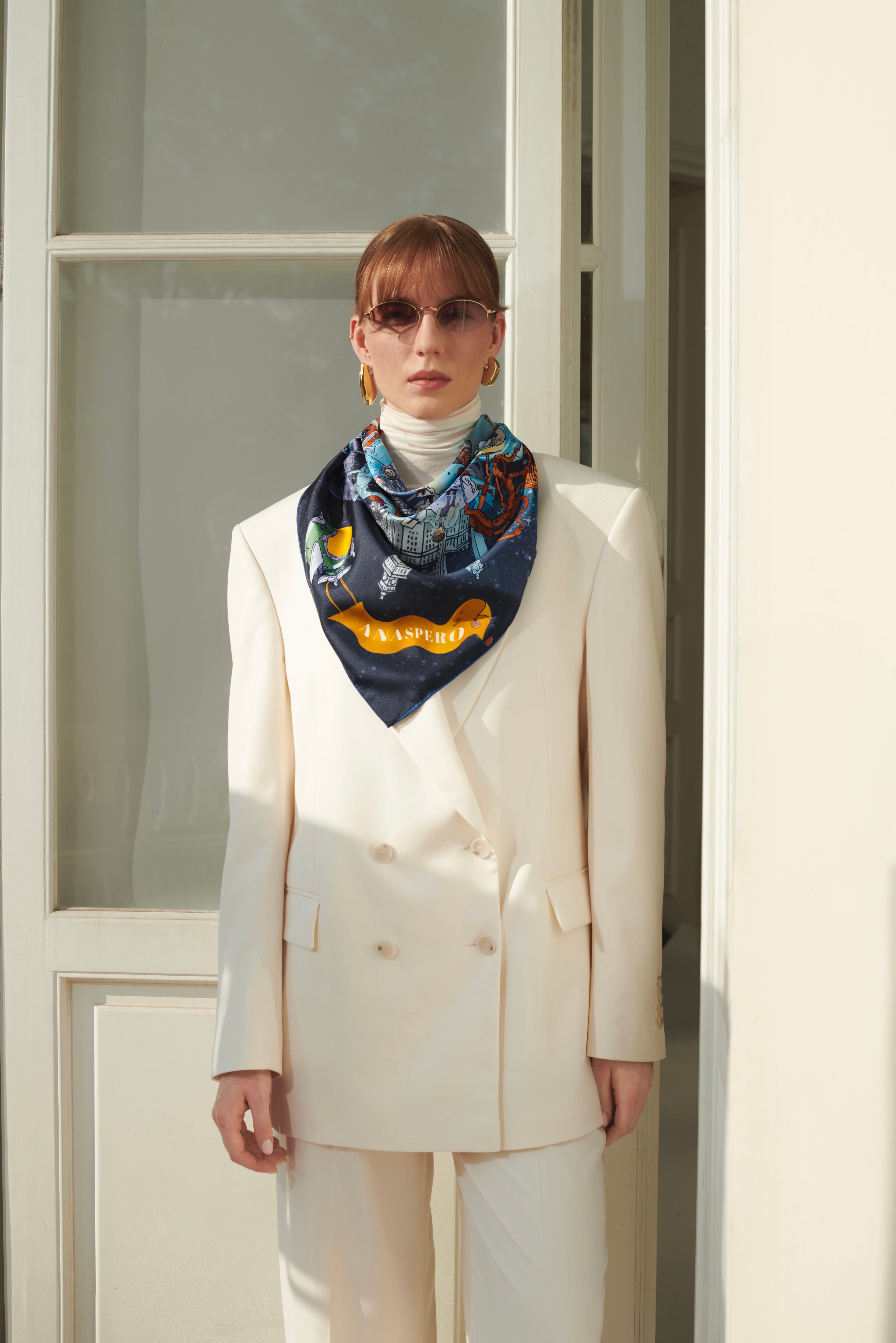 Person wearing a white suit and colorful scarf standing in front of a white door.